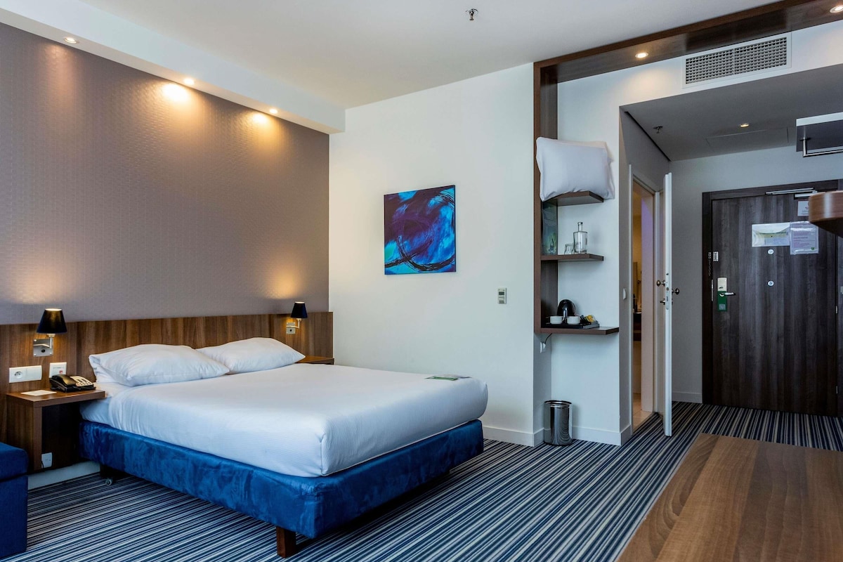 A hotel room with a bed, bedside table, lamp, artwork, and a door leading to a bathroom.
