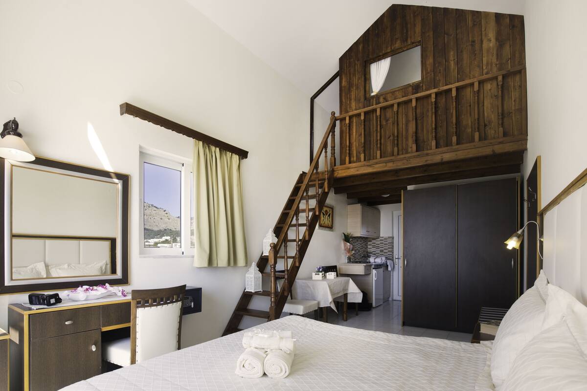 A bedroom with a bed, a desk, a mirror, a wardrobe, a staircase, and a small kitchen area.