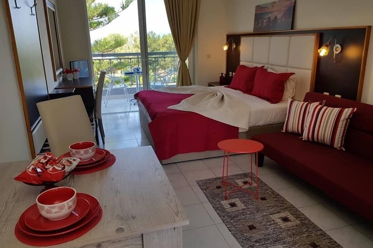 A hotel room with a bed, a sofa, a dining table with red tableware, and a balcony with chairs.