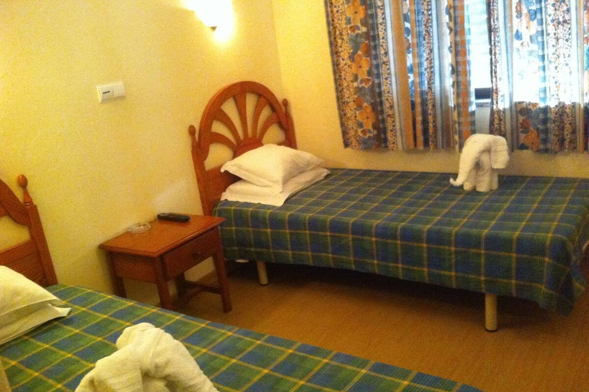 Quarto de hotel com duas camas de solteiro, uma mesinha de cabeceira de madeira, uma cadeira e janela com cortinas florais.