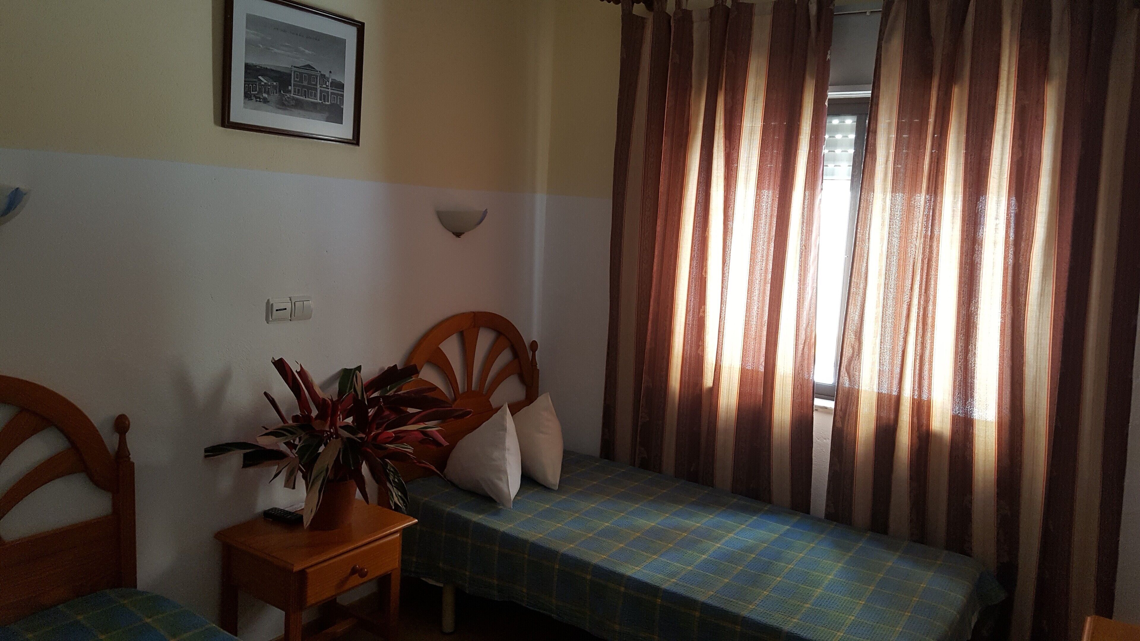 Quarto com duas camas de solteiro, uma mesinha com uma planta, uma luminária na parede e um quadro emoldurado.