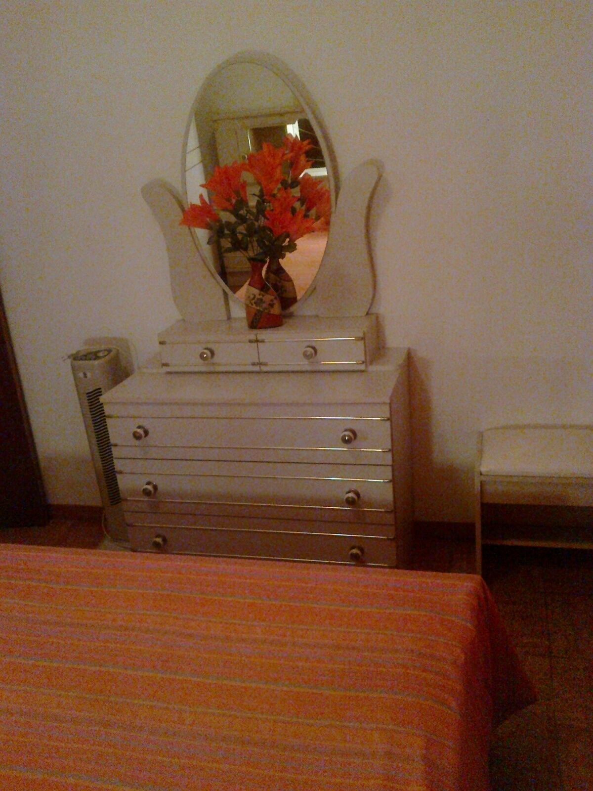 A room with a bed, a dresser with drawers, a mirror, and a vase with flowers on the dresser.