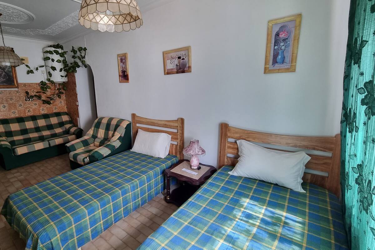 Quarto com duas camas, um sofá e uma mesa pequena. Há quadros nas paredes e um ventilador de teto.