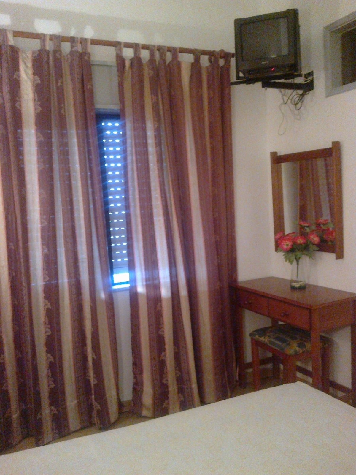 A room with a bed, a desk with a chair, a television mounted on the wall, and a window with curtains.
