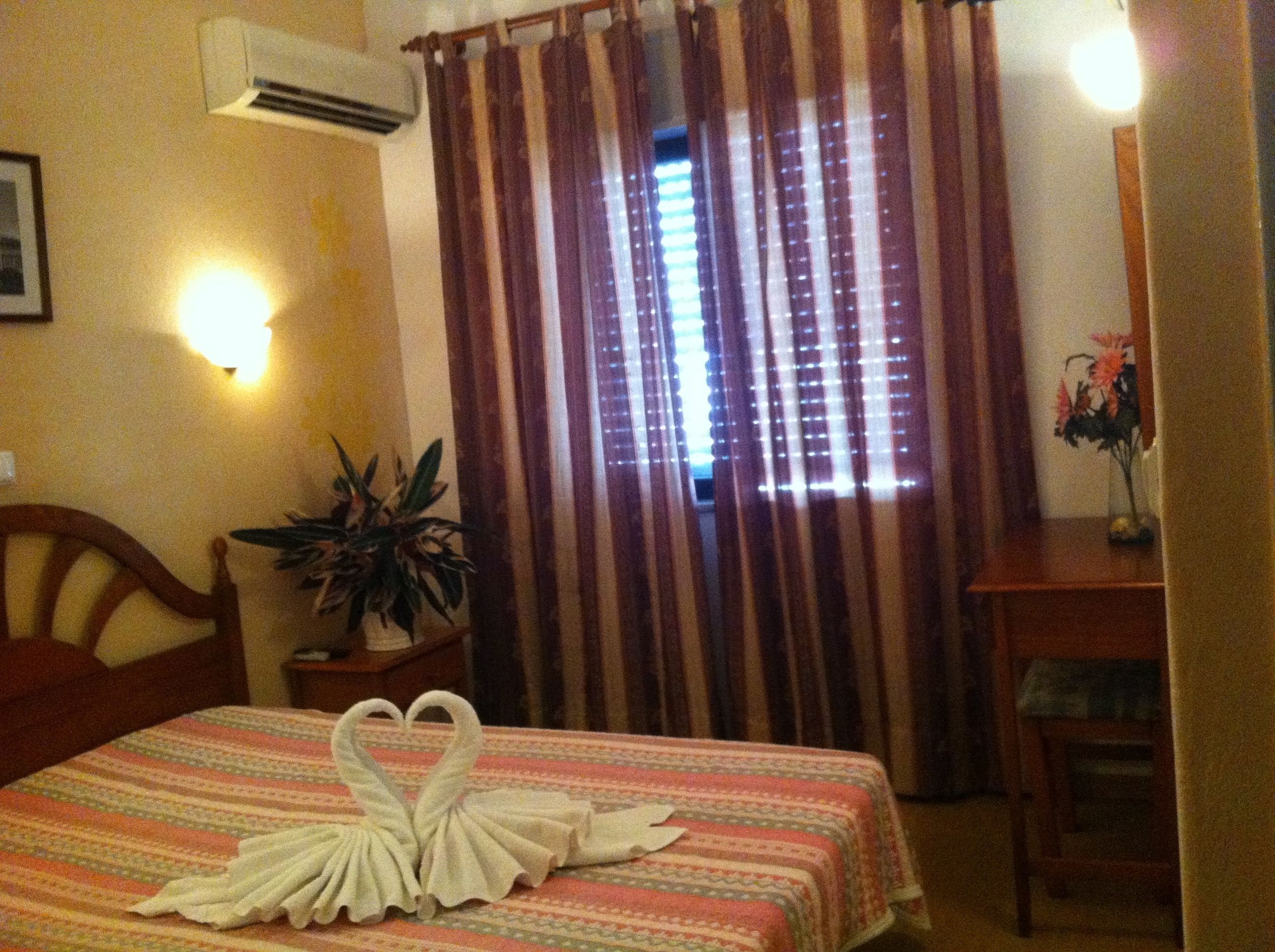 A hotel room with a bed, a bedside table with a plant, a wall-mounted air conditioner, and a window with curtains.