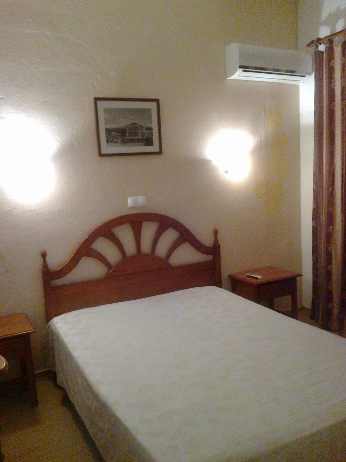 A bedroom with a wooden bed, a bedside table, a nightstand, a framed picture on the wall, and an air conditioning unit.