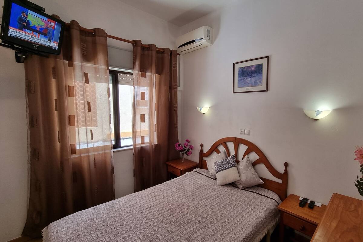 Quarto com cama, mesa de cabeceira, televisão, ar-condicionado, quadro emoldurado e janela com cortinas.