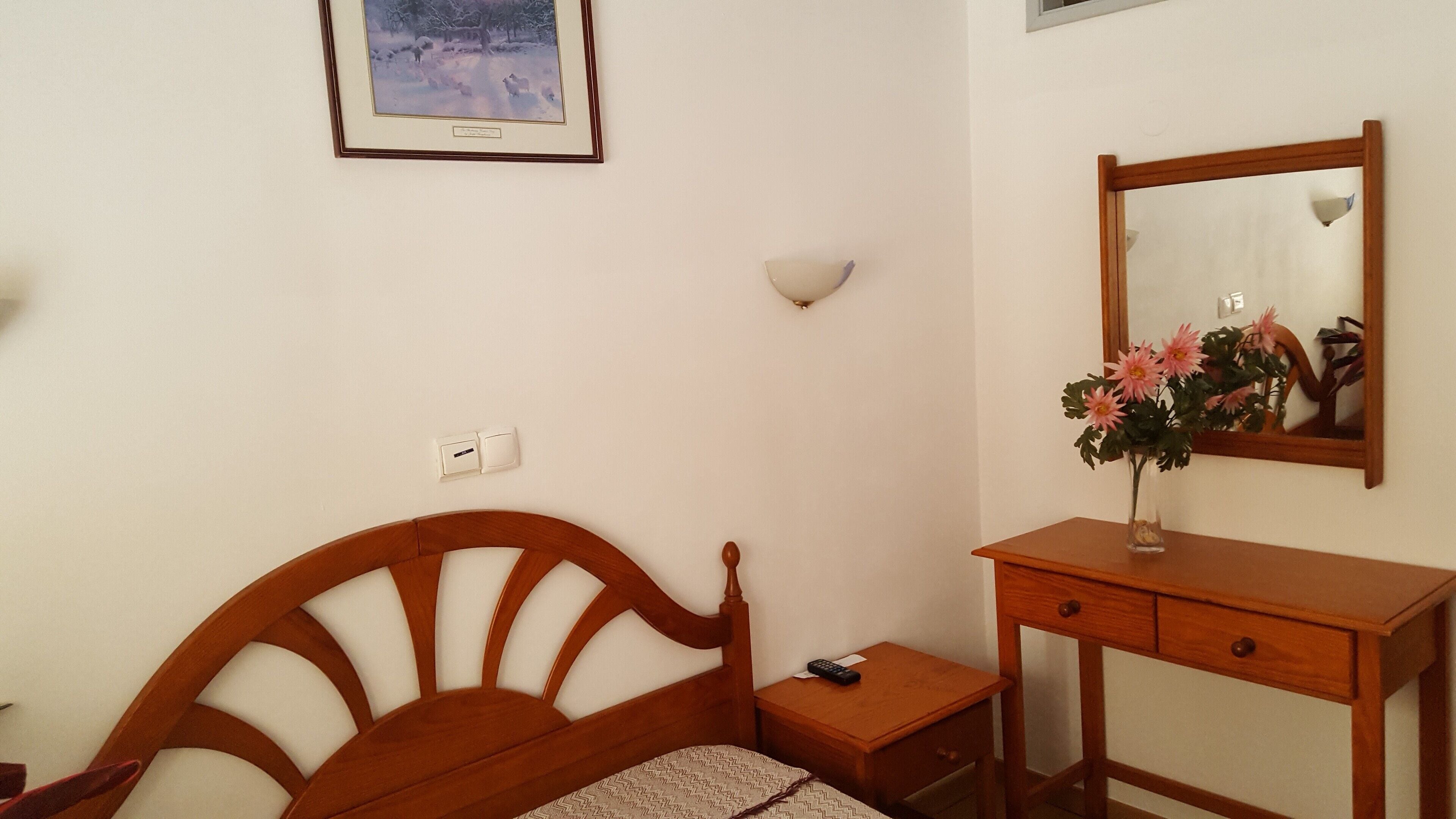 Ein Schlafzimmer mit einem hölzernen Bett, einem Nachttisch, einem kleinen Schreibtisch mit Spiegel und einem gerahmten Bild an der Wand.