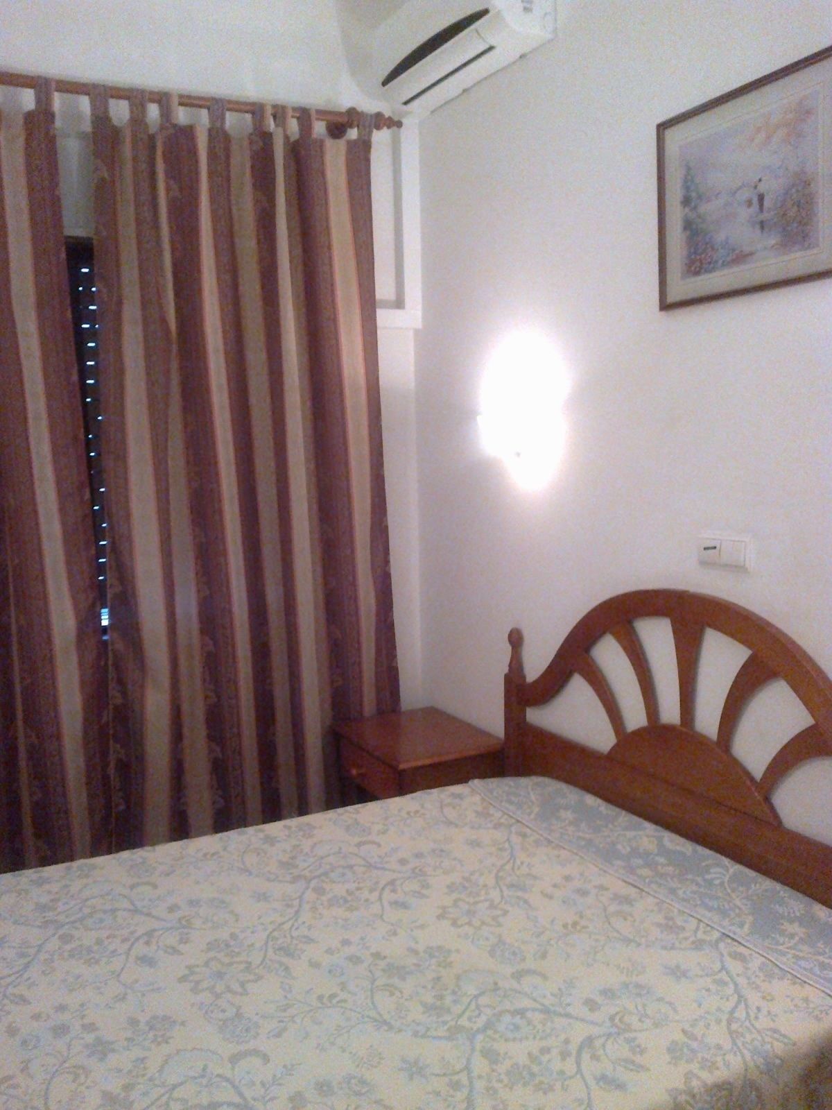 Un dormitorio con cama, mesita de noche, luz de pared, ventana con cortinas y un cuadro en la pared.