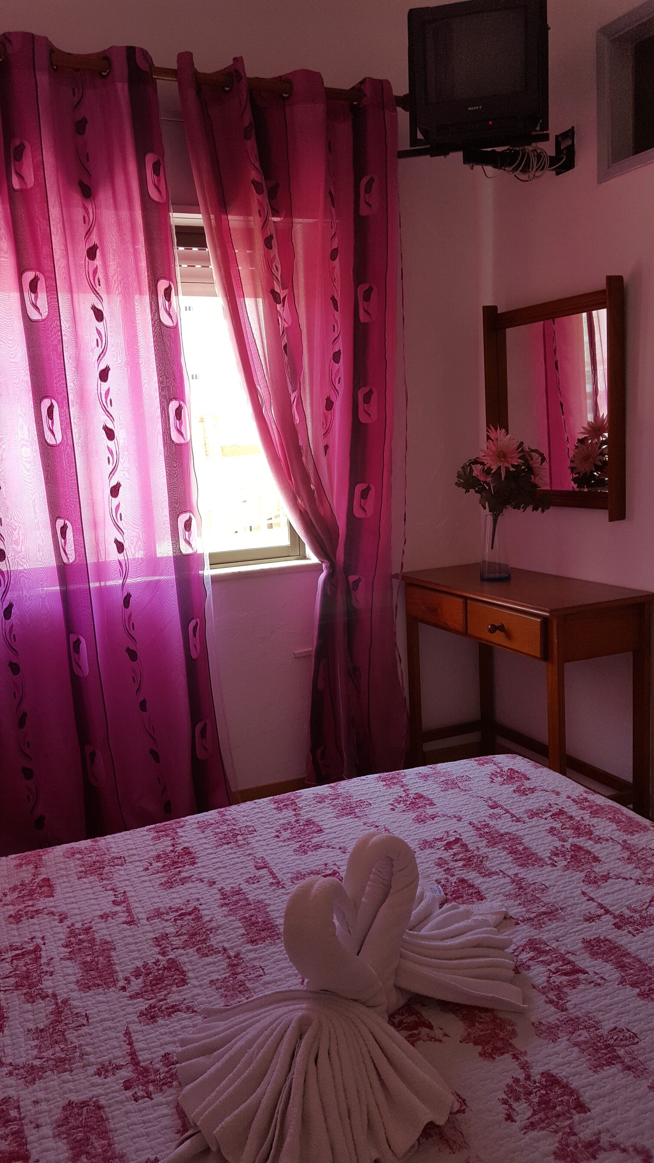 Une chambre d’hôtel avec un lit, une table de chevet en bois, un miroir et des rideaux roses.