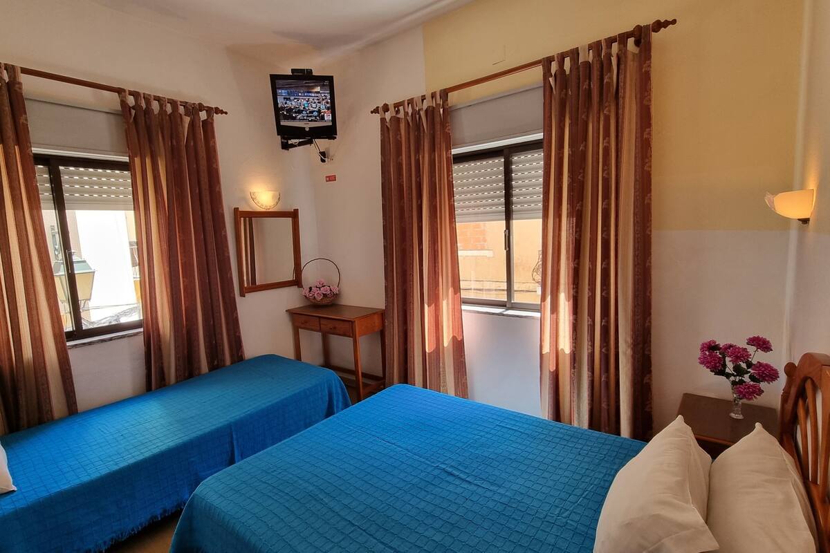 Quarto de hotel com duas camas de solteiro, uma televisão na parede, uma mesinha com abajur e uma janela com persianas.