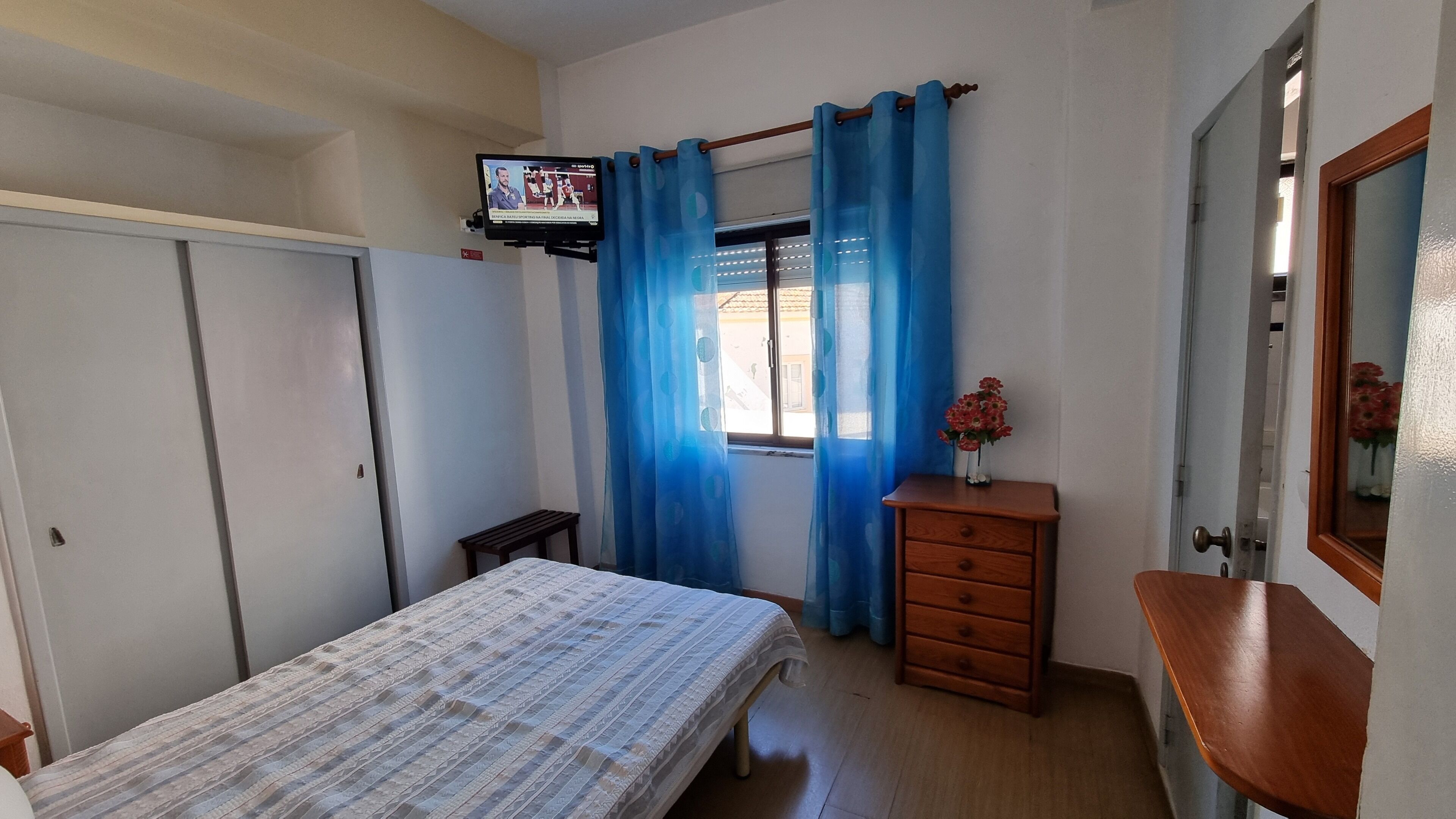 A bedroom with a bed, a dresser, a small table, a TV mounted on the wall, and blue curtains.