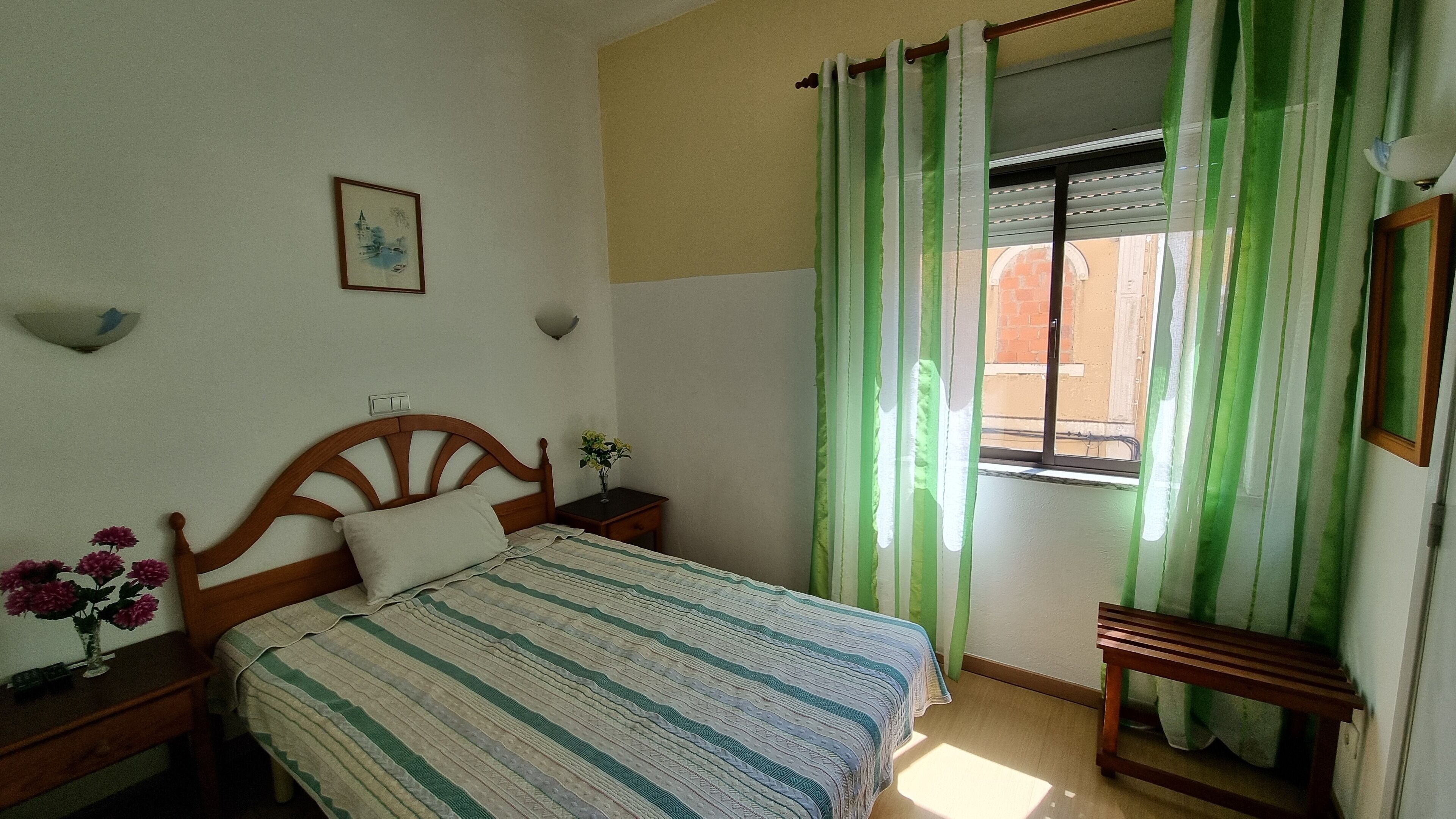 A bedroom with a bed, a nightstand, a bedside table, a window with green curtains, and a wall-mounted light fixture.