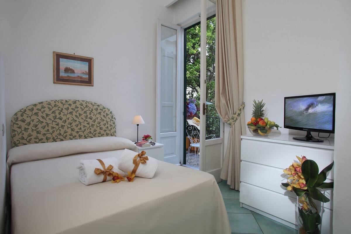 Un lit bien fait, avec une serviette et un coussin décoratif, une télévision posée sur une commode, et une fenêtre avec des rideaux.
