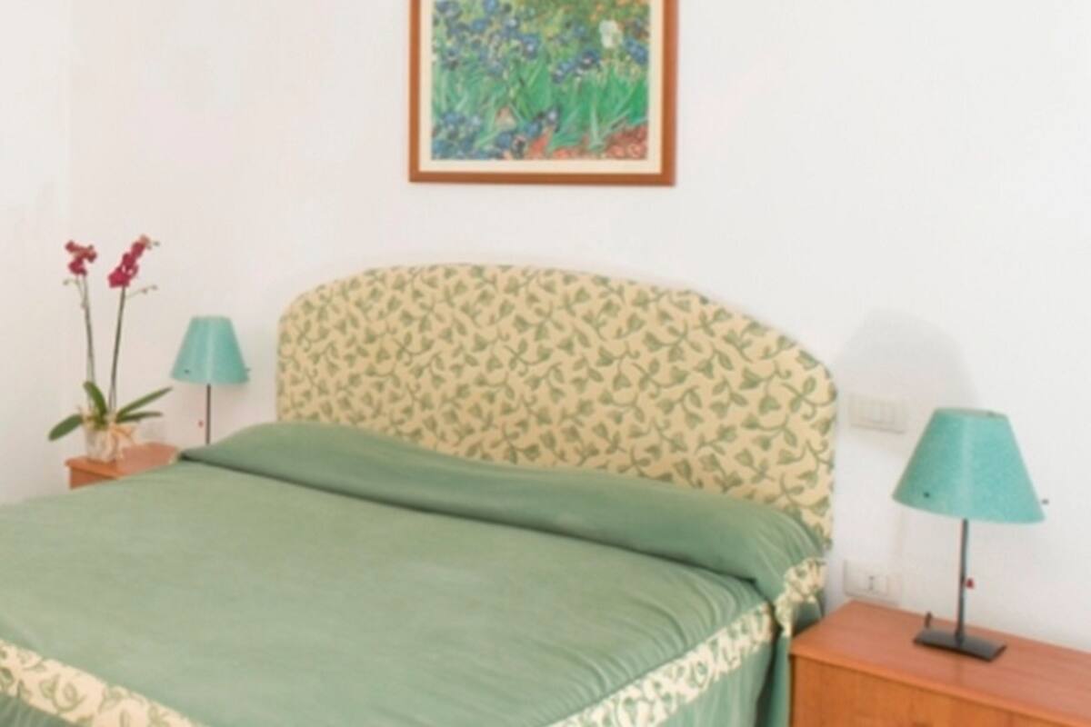 Une chambre avec une tête de lit à motifs, une couvre-lit verte, deux lampes de chevet et un tableau floral encadré accroché au mur.