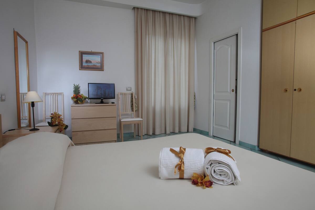 Une chambre d’hôtel comprenant un lit, un bureau avec un ordinateur, une télévision, une armoire et une fenêtre avec des rideaux.