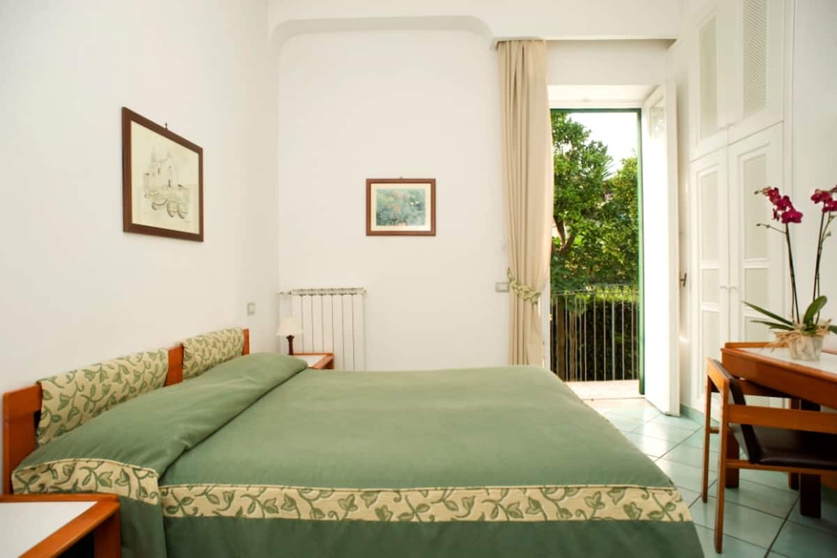 Une chambre avec un grand lit, une petite table et une chaise. Il y a une fenêtre avec des rideaux et une vue sur la verdure à l’extérieur.