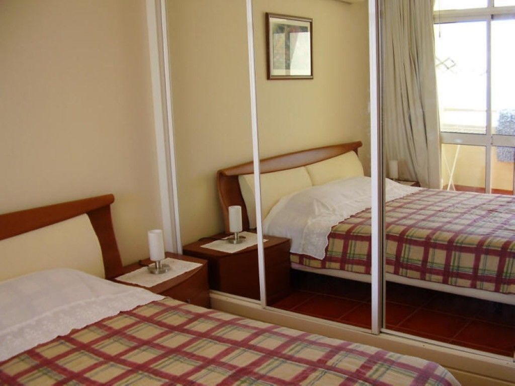 A hotel room with two beds, a mirror, a nightstand with a lamp, and a window with curtains.
