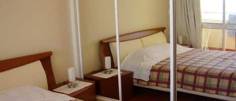A hotel room with two beds, a mirror, a nightstand with a lamp, and a window with curtains.