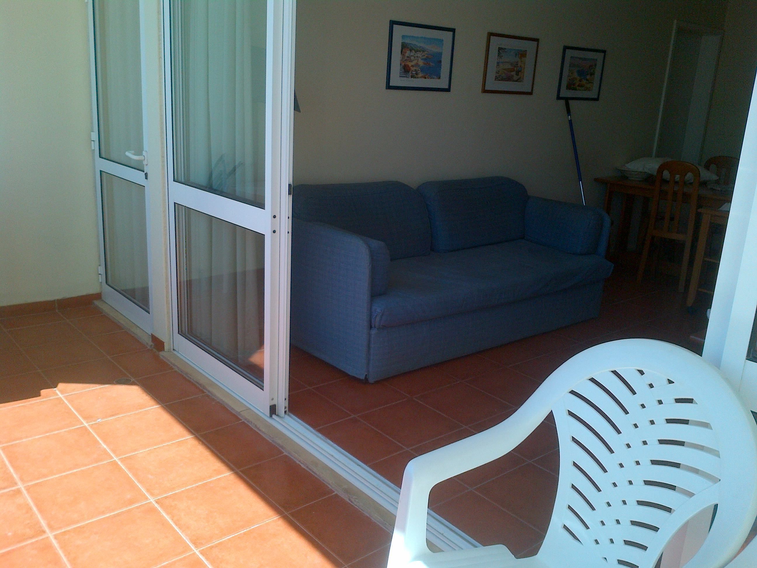A living area with a blue sofa, a white chair, and a sliding glass door leading to an outdoor space.