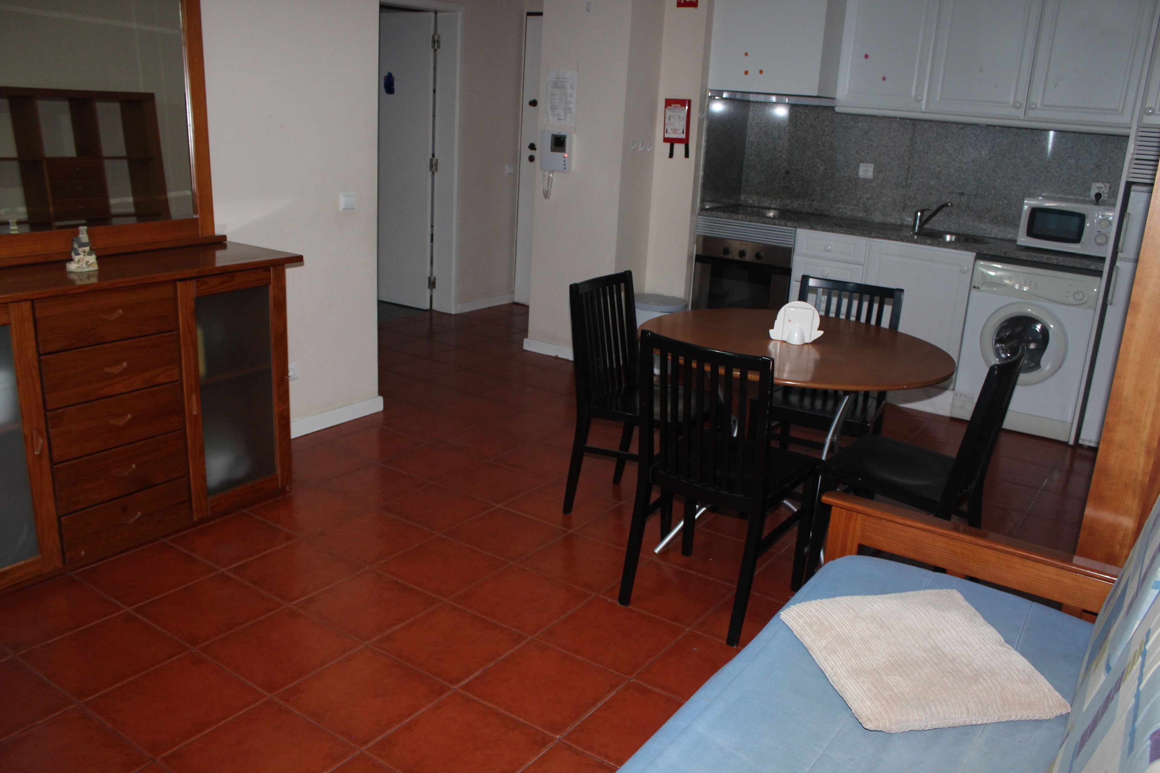 A compact kitchen with a microwave, oven, and washing machine. There is a dining table with chairs and a small wooden dresser with drawers.