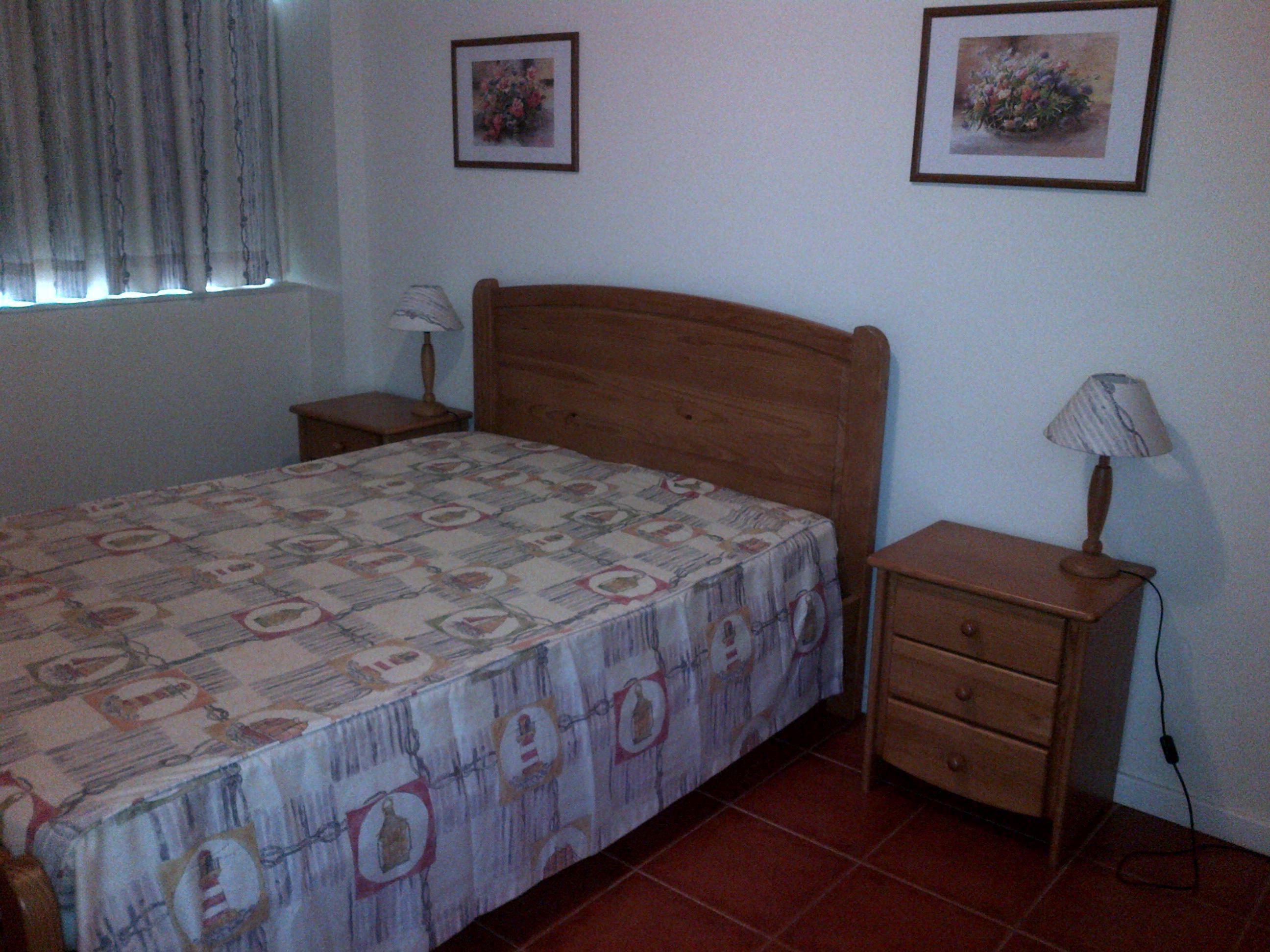 A bedroom with a bed, two nightstands, a wooden headboard, a window with curtains, and two framed pictures on the wall.