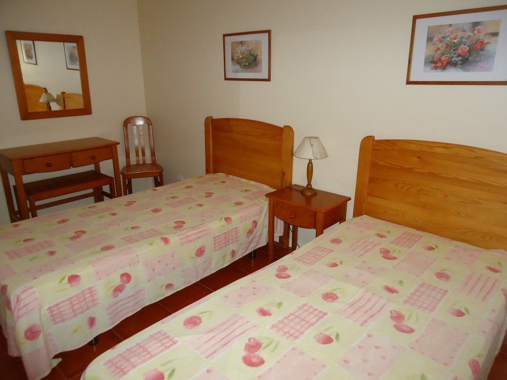 A room with two single beds, a wooden desk, a chair, and a small bedside table with a lamp.