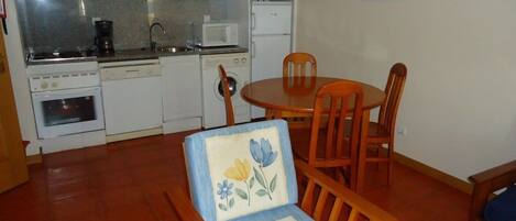 Une cuisine de taille réduite avec des appareils électroménagers blancs, une table à manger avec quatre chaises et un canapé simple recouvert d’un tissu à motifs floraux.