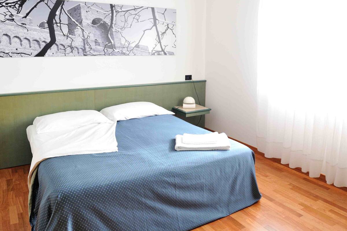 A neatly made bed with a blue bedspread and white pillows, a bedside table with a lamp, and a wall with a black and white artwork.