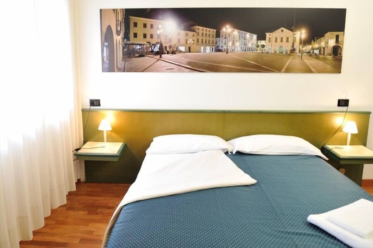 A hotel room with a large bed, two bedside lamps, and a cityscape view on the wall.