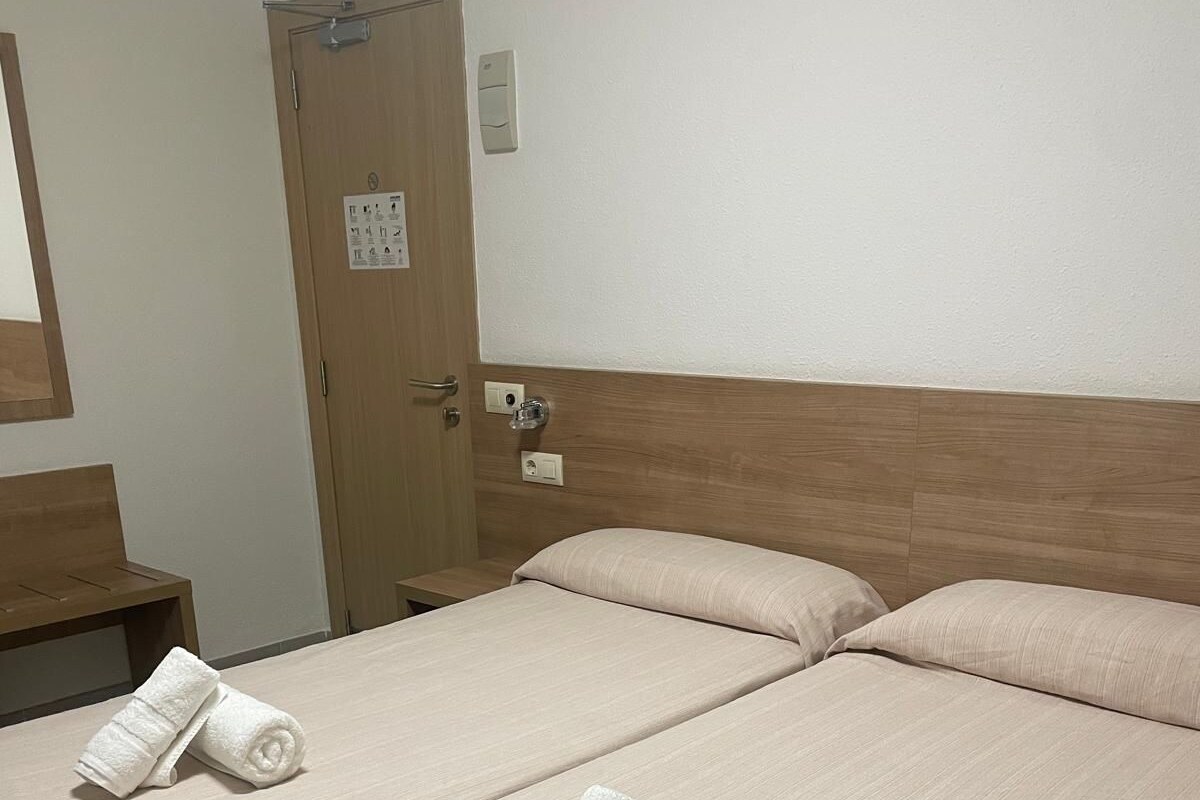 Habitación de hotel con dos camas, cada una con cubrecama color beige y dos toallas blancas enrolladas. Se observa un cabecero de madera, un banco de madera y una puerta con un cartel encima.