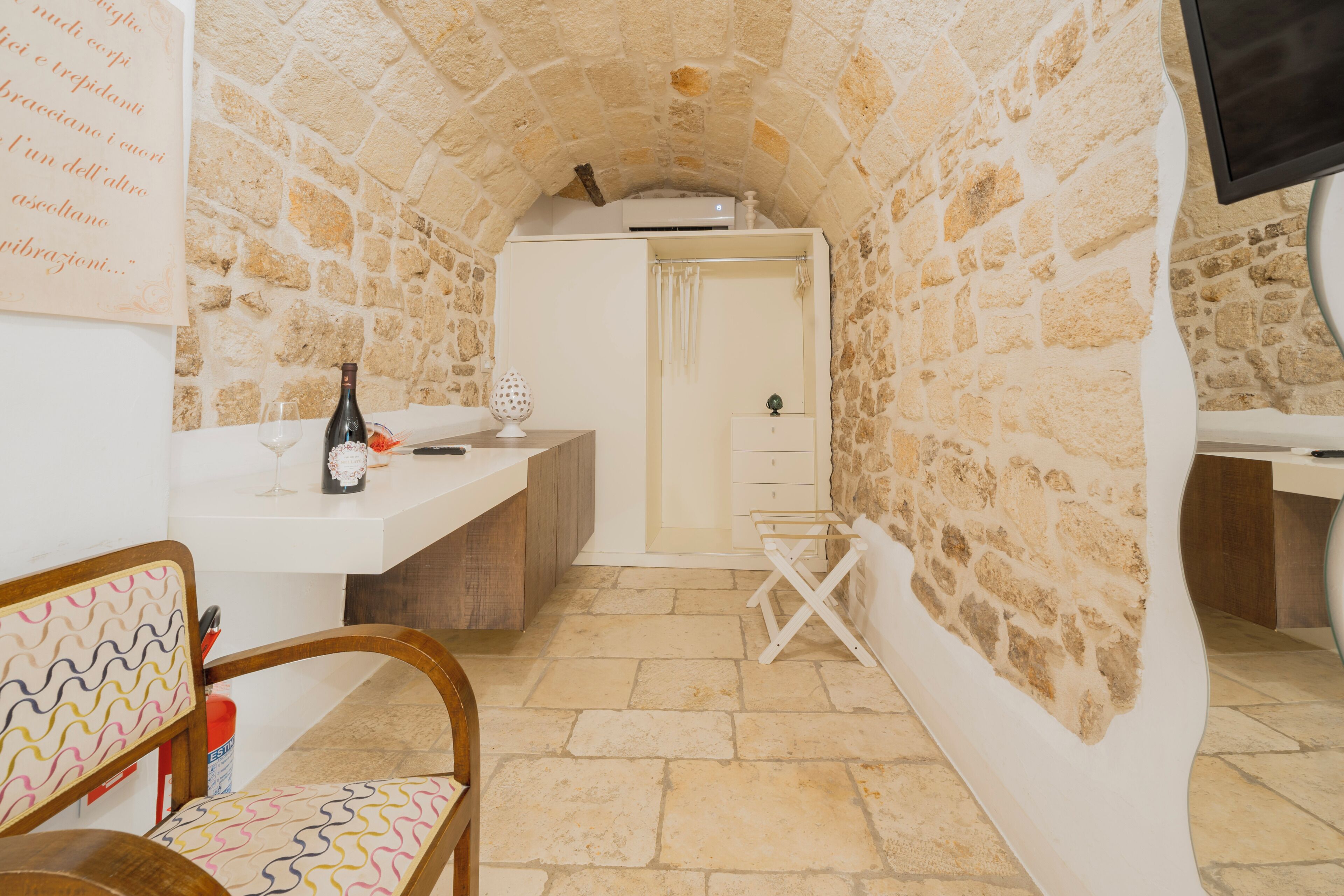 Habitación con paredes de piedra, un lavamanos blanco, una silla de madera, una botella y una copa de vino sobre una encimera.