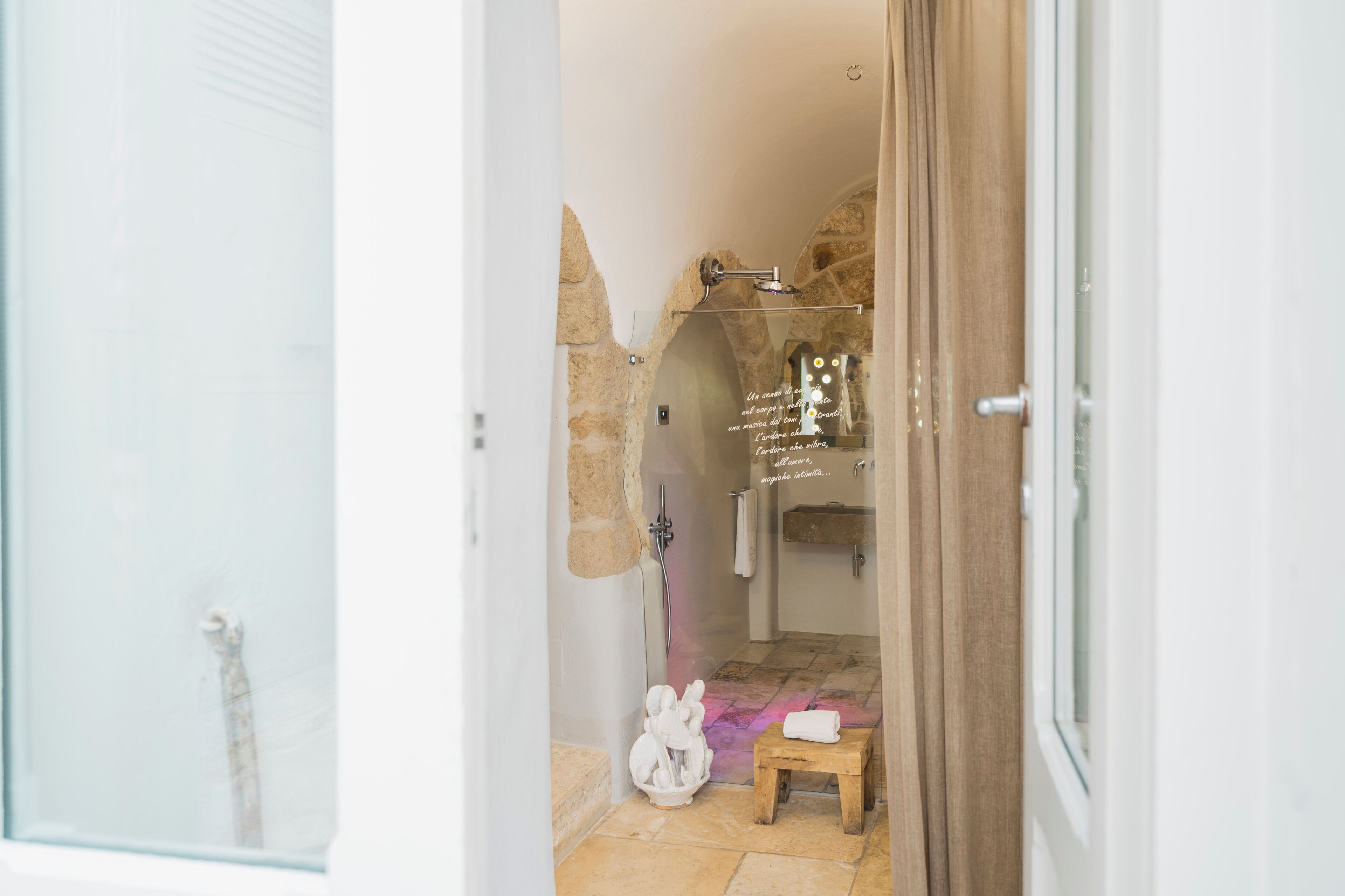 A bathroom with a stone arch, a shower, a small wooden stool, and a towel on the floor.