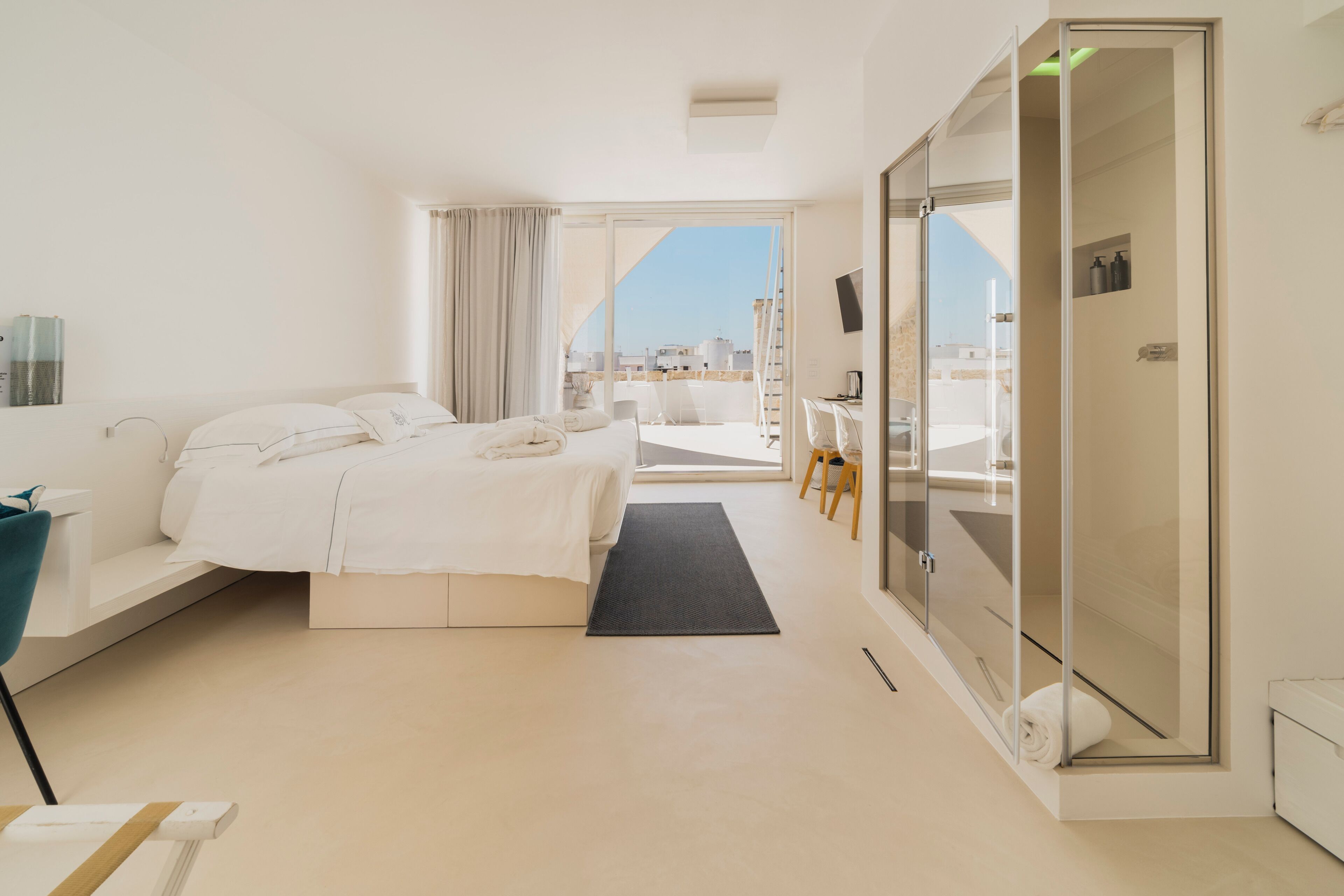 A modern hotel room with a large bed, a desk, a chair, a glass shower enclosure, and a view of the city.