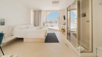 A modern hotel room with a large bed, a desk, a chair, a glass shower enclosure, and a view of the city.