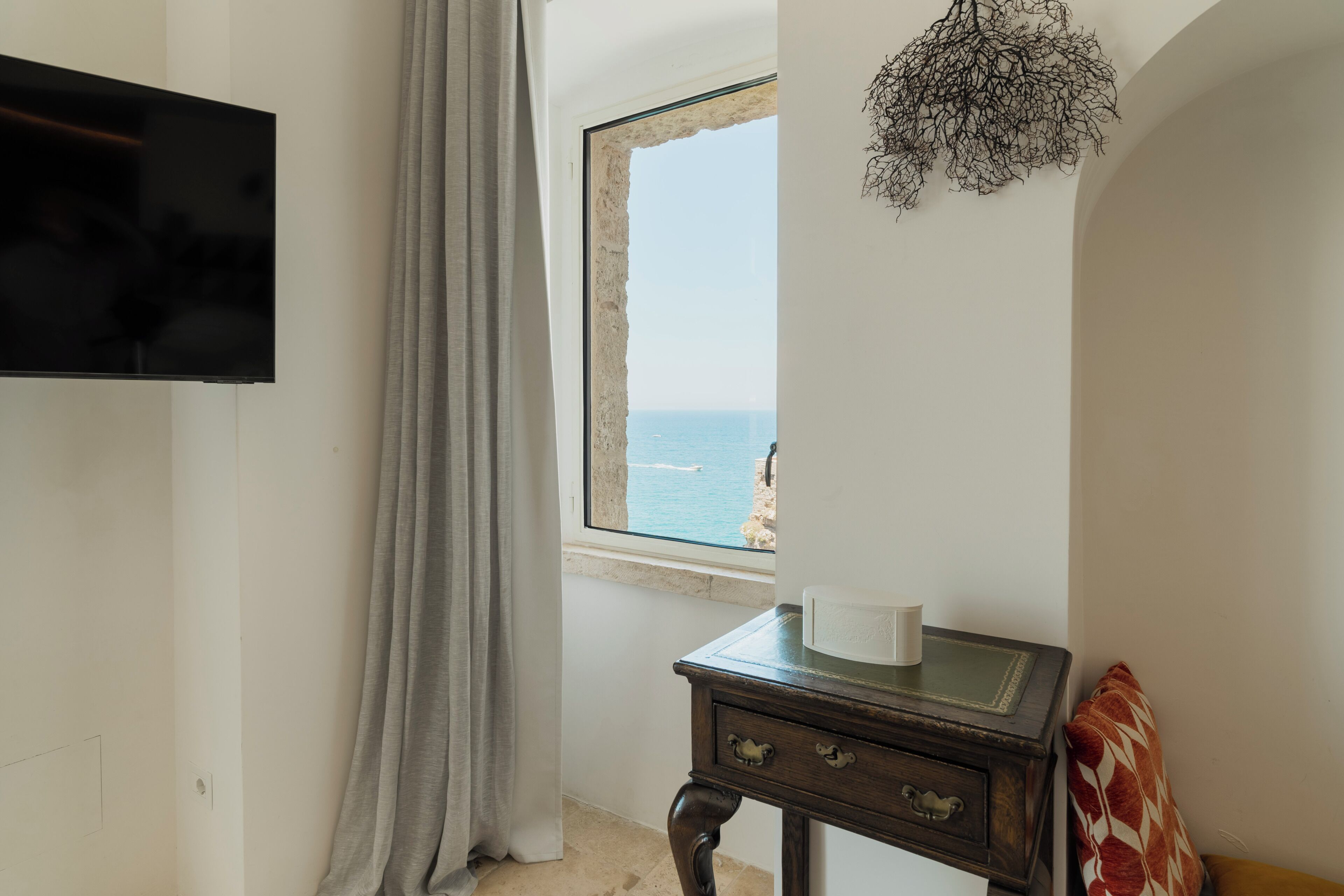 A room with a TV, a window with a view of the sea, a small table with a cup, and a decorative wall piece.