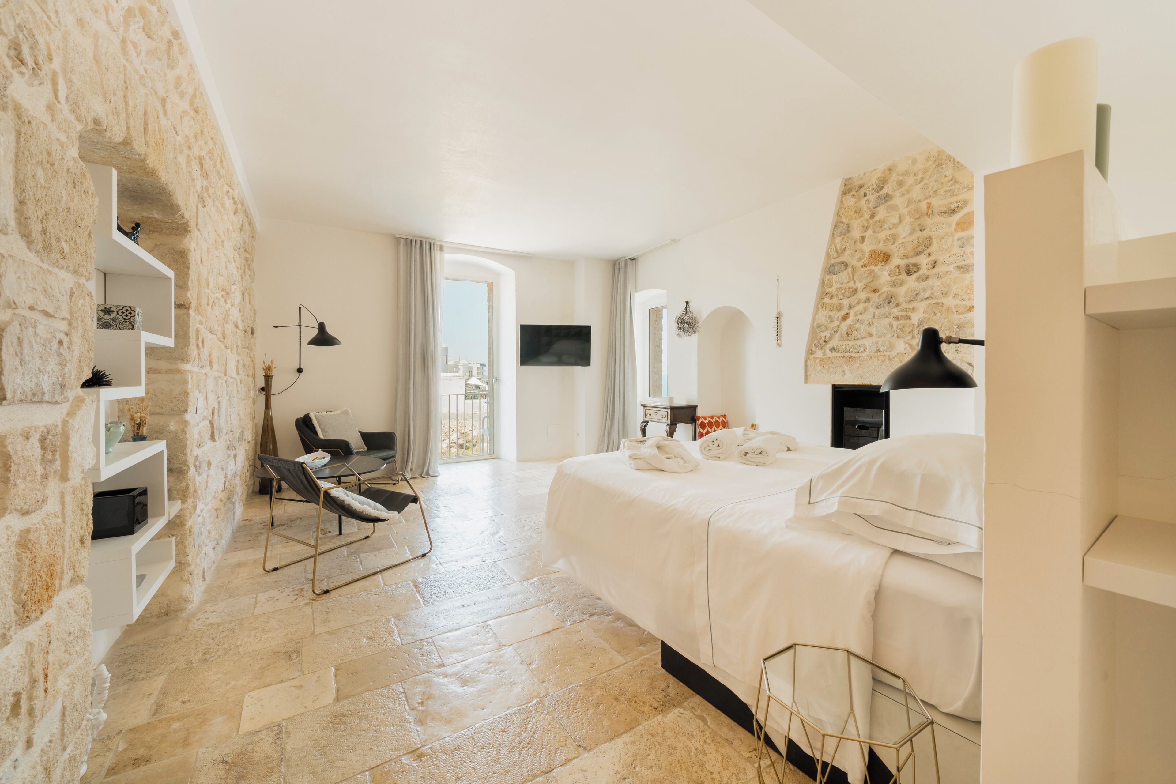 A hotel room with stone walls, a bed, a chair, a desk, and a view of the city.