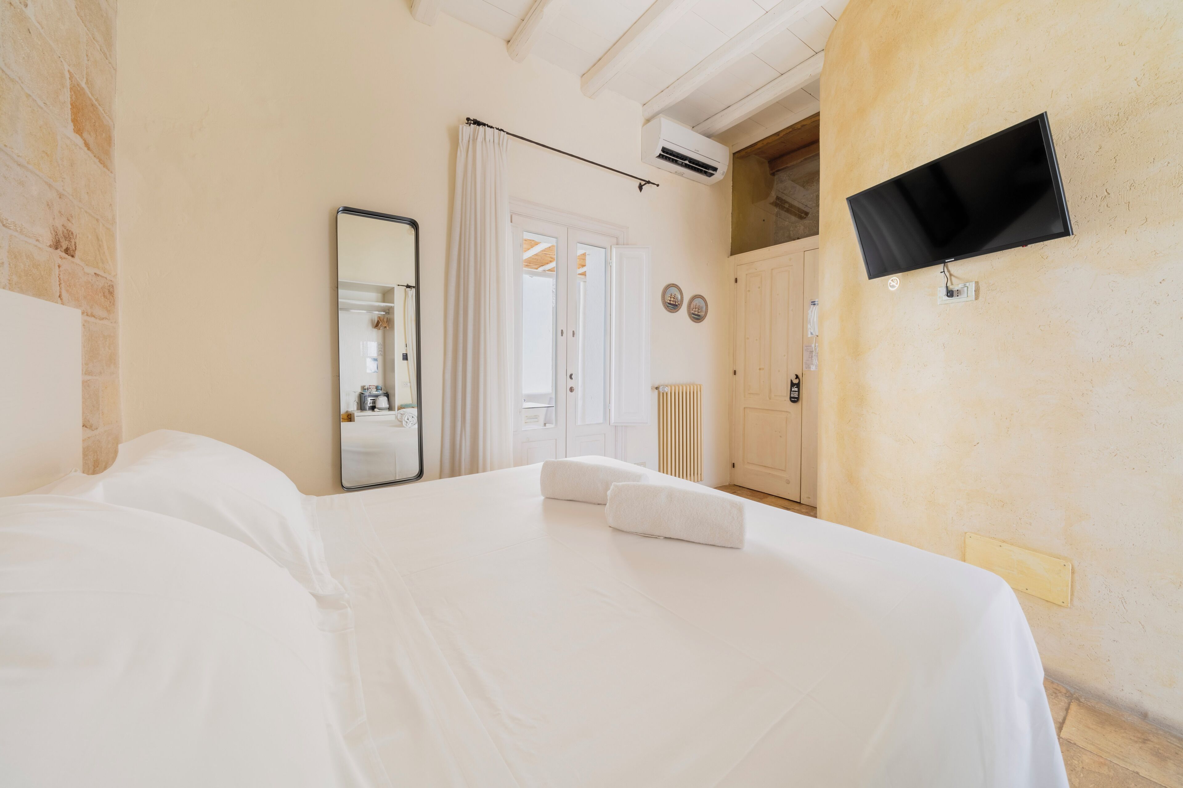 Un dormitorio con cama, espejo, televisor y puerta que da a otra habitación.