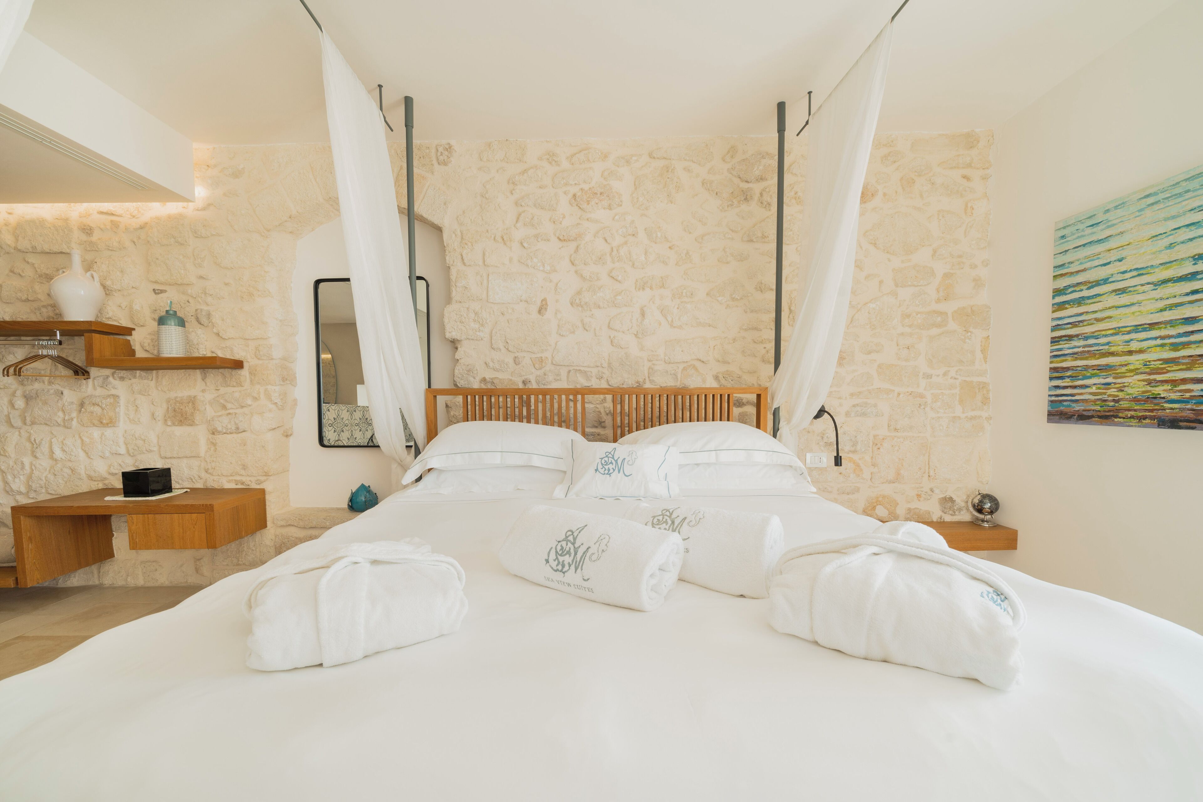 A bedroom with a stone wall, wooden bed frame, white bedding, and a painting on the wall.