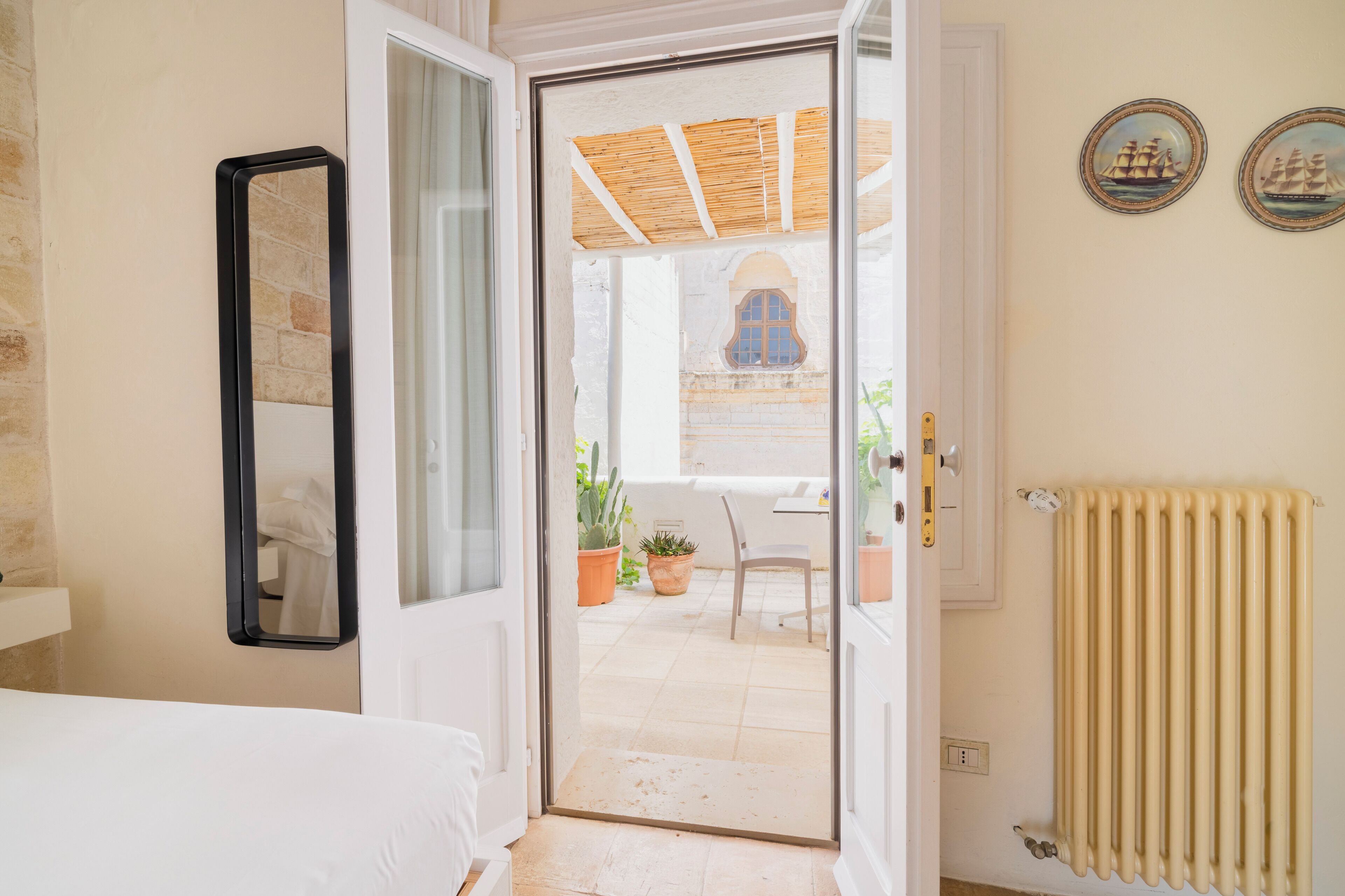Une chambre d’hôtel avec un lit, une porte donnant sur un patio extérieur agrémenté de plantes et d’une chaise, un radiateur et deux tableaux encadrés au mur.