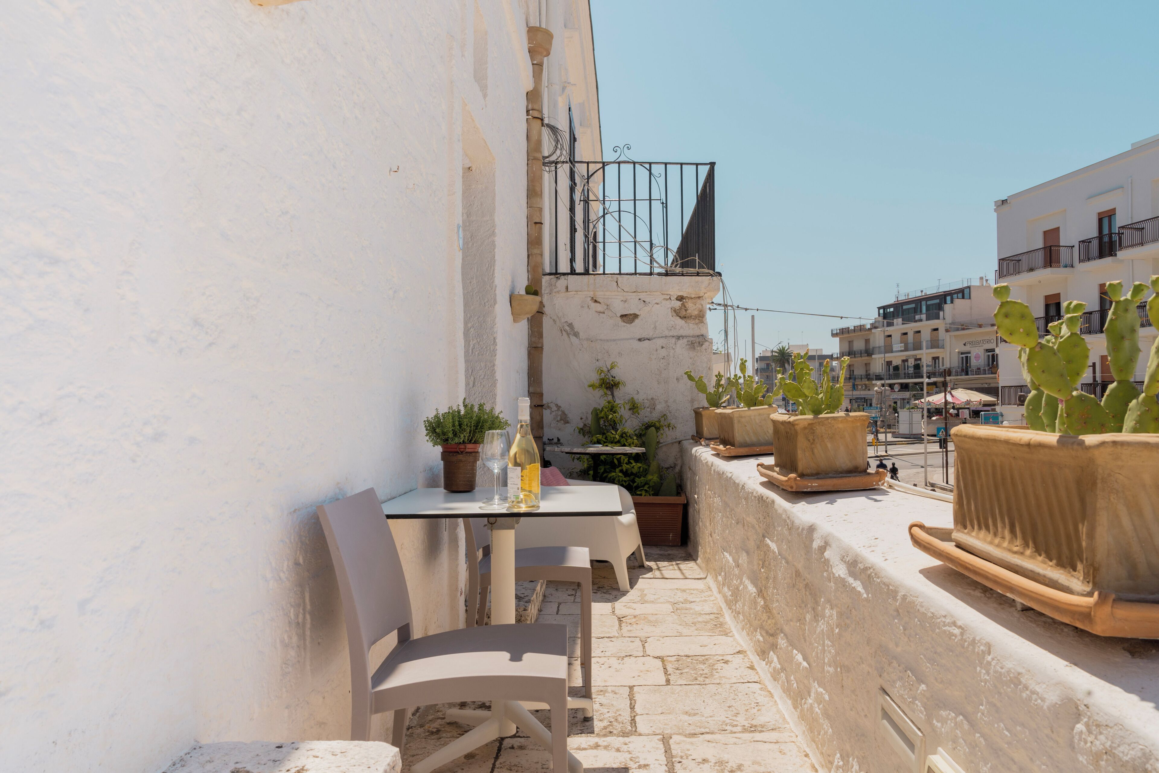 A narrow outdoor balcony with a table and chairs, potted plants, and a view of the street and buildings.