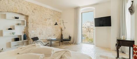 A hotel room with a stone wall, a bed with towels, a desk with a chair, a TV, and a balcony with a view.