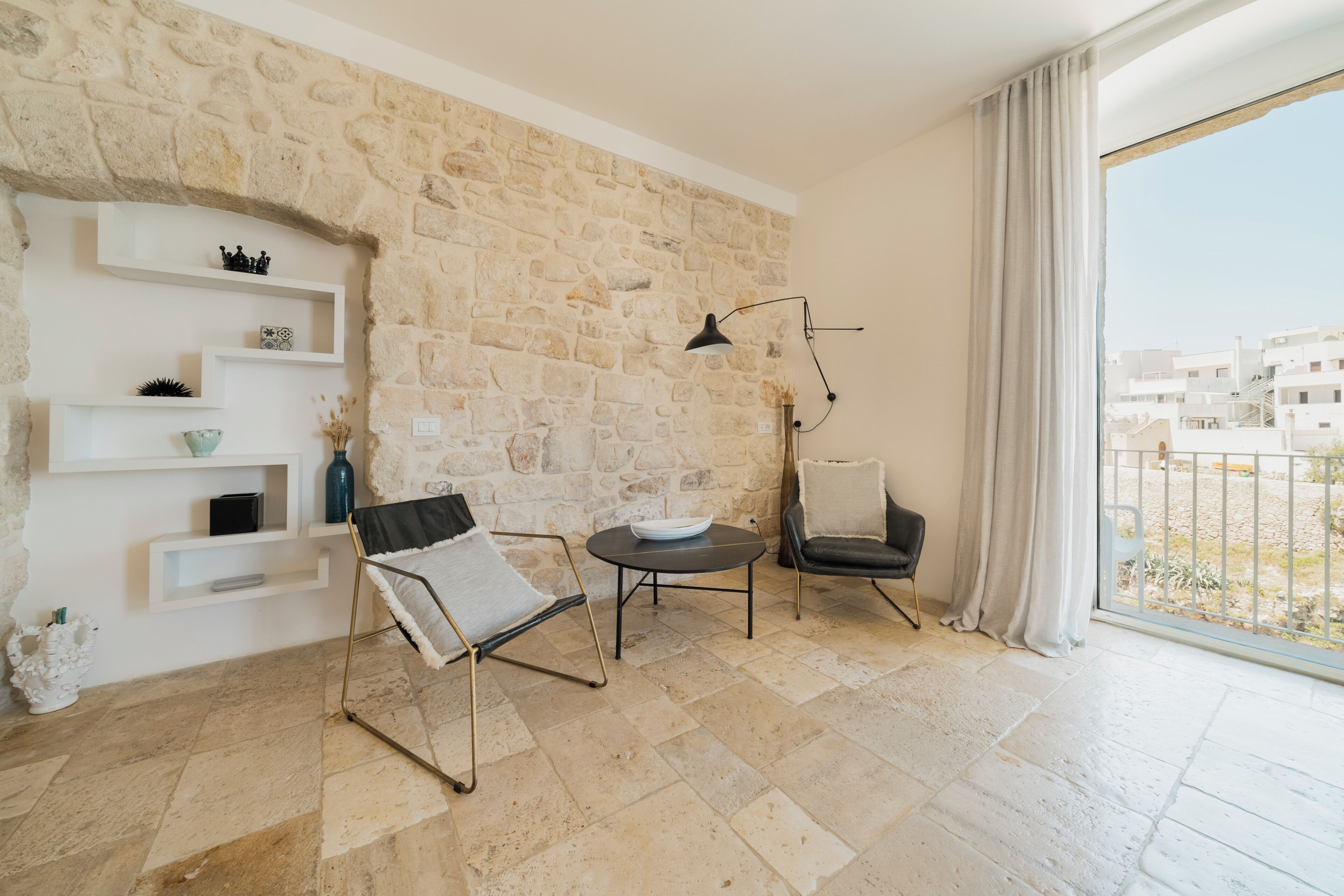 A modern living room with a stone wall, a black floor lamp, a round table, a black chair, and a view of buildings through a large window.