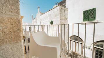 Un balcón con una silla blanca, un edificio blanco con contraventanas verdes y un cielo azul despejado.