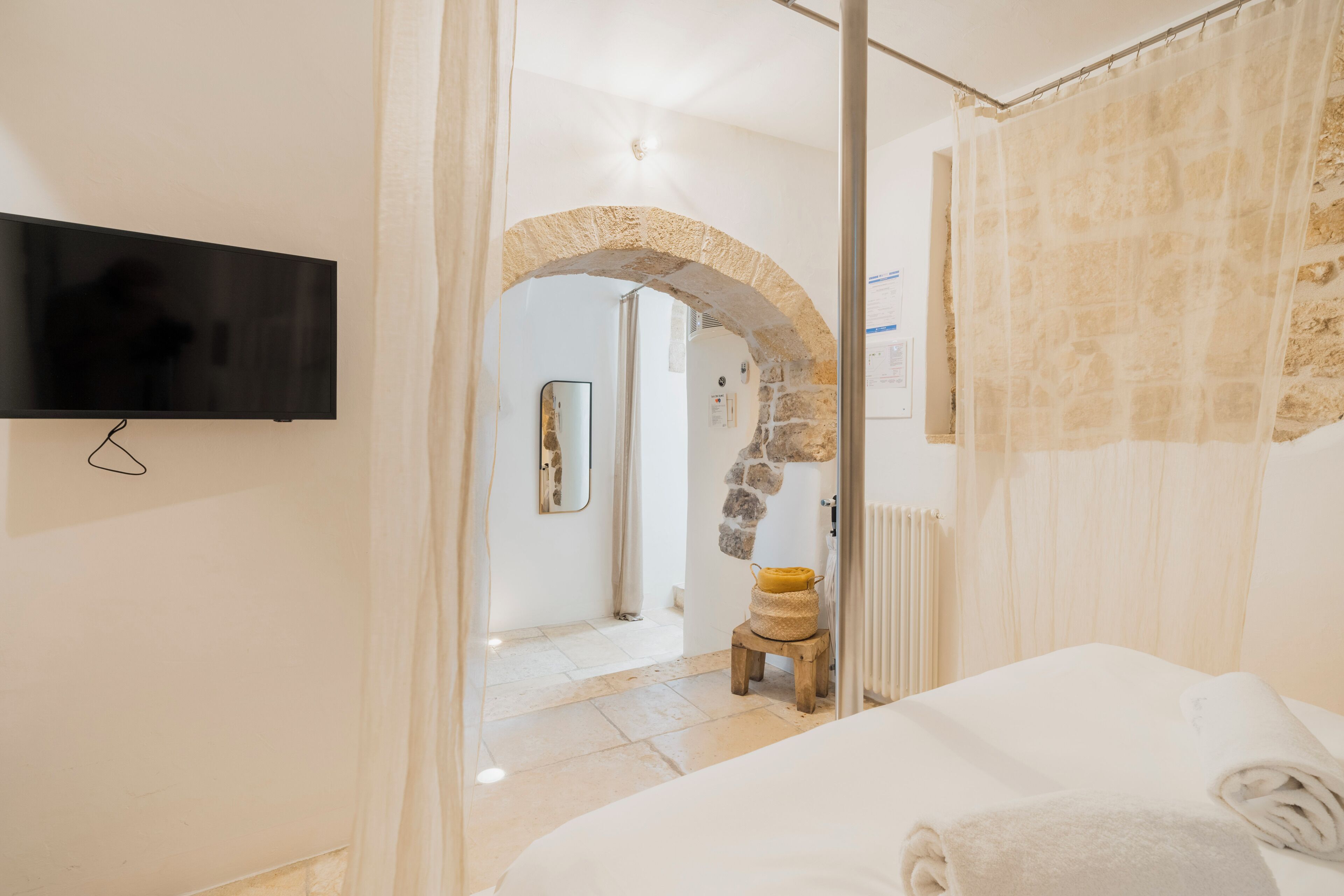 Une chambre à coucher avec un lit, un téléviseur fixé au mur, une arche en pierre et un petit tabouret.