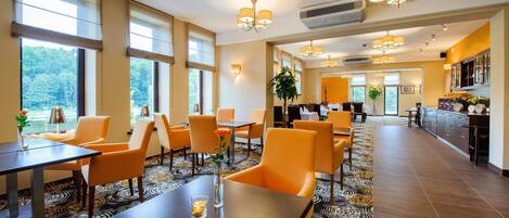 Een restaurantgedeelte in een hotel met oranje stoelen, een lange tafel en uitzicht op het bos door grote ramen.