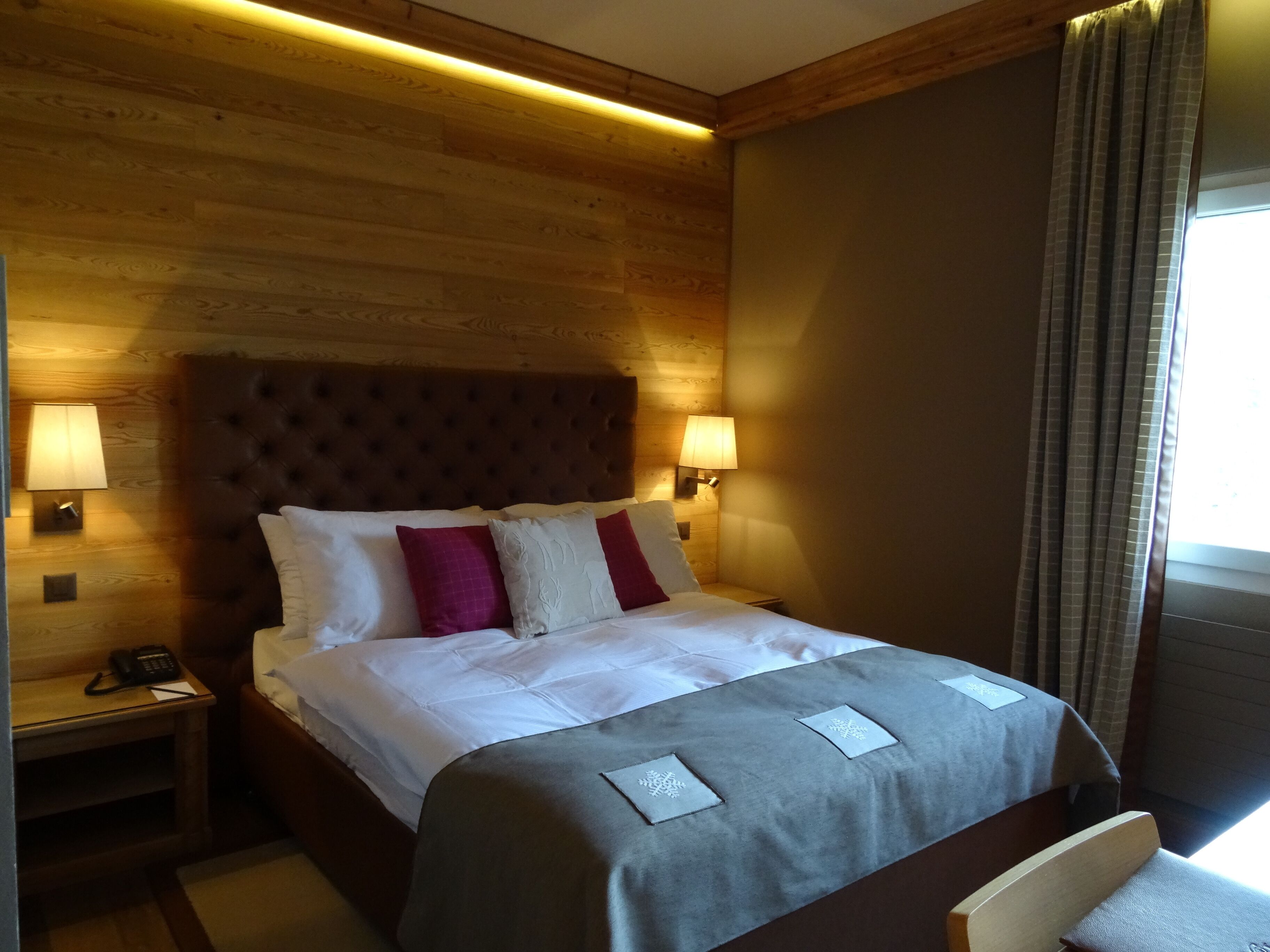 Habitación de hotel con cabecera de madera, dos lámparas de noche, un teléfono y una ventana con cortinas.