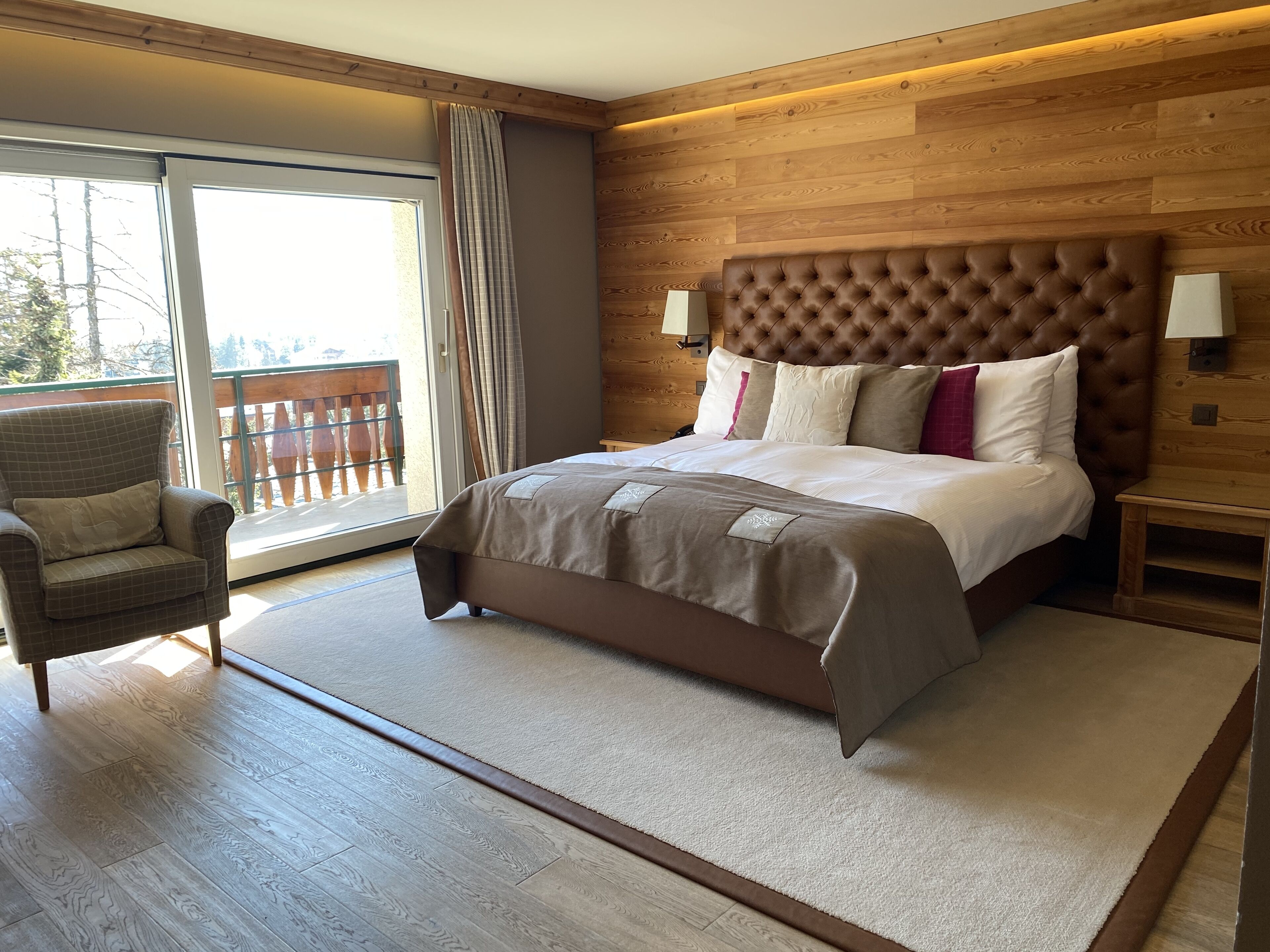 Ein Schlafzimmer mit einem großen Bett, einem Sessel, einem Holz-Kopfteil und Ausblick ins Freie durch eine Schiebetür.