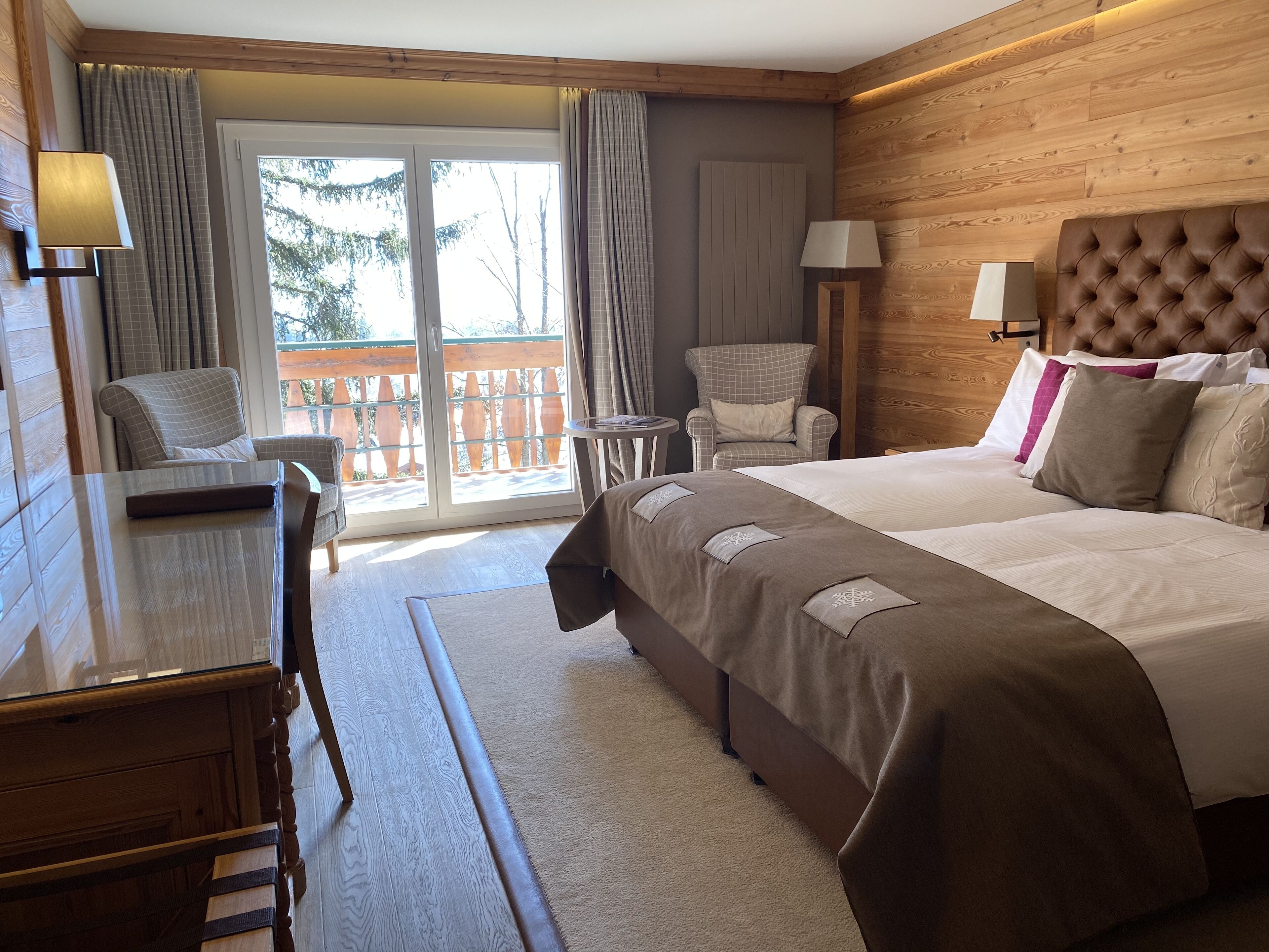 A hotel room with a large bed, a desk, a chair, and a view of the outdoors through a sliding glass door.