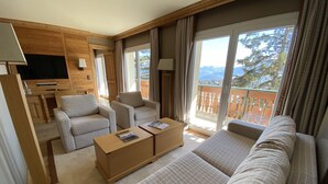 Habitación de hotel moderna con sofá, dos sillones, una mesa de centro y televisor. La habitación tiene ventanas amplias con vistas a montañas y un balcón.